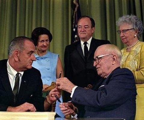 LBJ prepares to sign Medicare into law. Former President Harry Truman hands him the pen, as VP Hubert Humphrey, Lady Bird Johnson and Bess Truman look on.