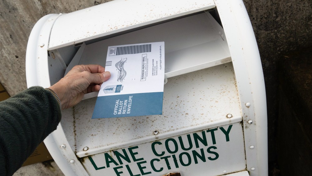 A hand hovers a ballot over a ballot drop box
