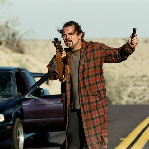 Leonardo Di Caprio stands in a robe on the street holding a gun.