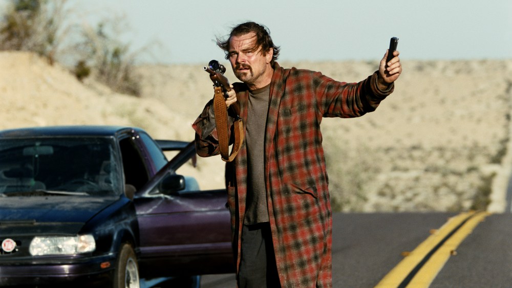 Leonardo Di Caprio stands in a robe on the street holding a gun.