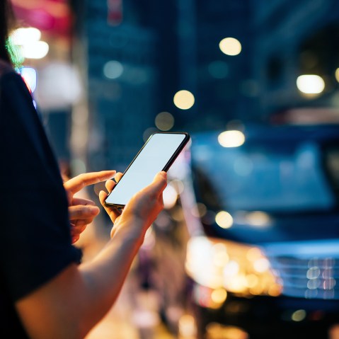 Close up of young woman using mobile app device on smartphone to hail a taxi ride on city street after work in the evening.