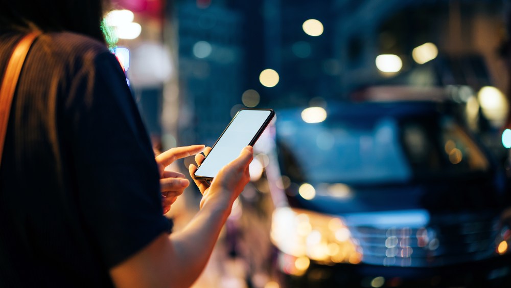 Close up of young woman using mobile app device on smartphone to hail a taxi ride on city street after work in the evening.