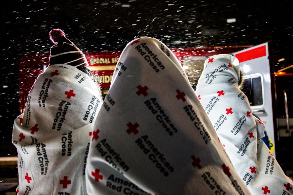 Three people bundled in American Red Cross blankets wait in the snow beside an American Red Cross truck.