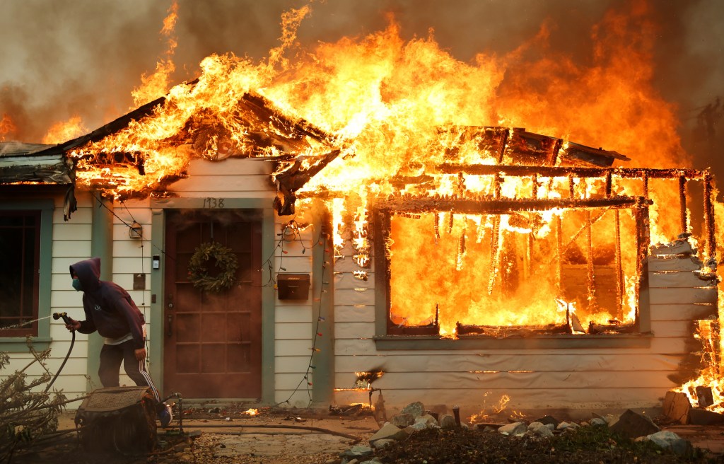 A person uses a garden hose to put out the flames of a house on fire.