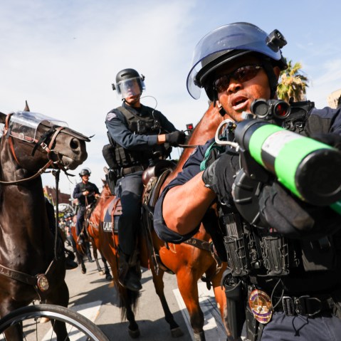 A police officer holds a weapon to the camera as protesters face off with police outside of a federal building in downtown Los Angeles for an anti-Trump "No Kings Day" demonstration.