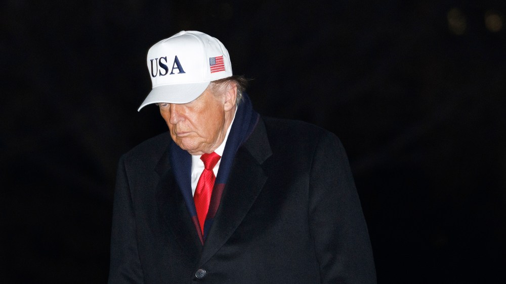 Donald Trump walks at the White House at night, wearing a white USA hat.