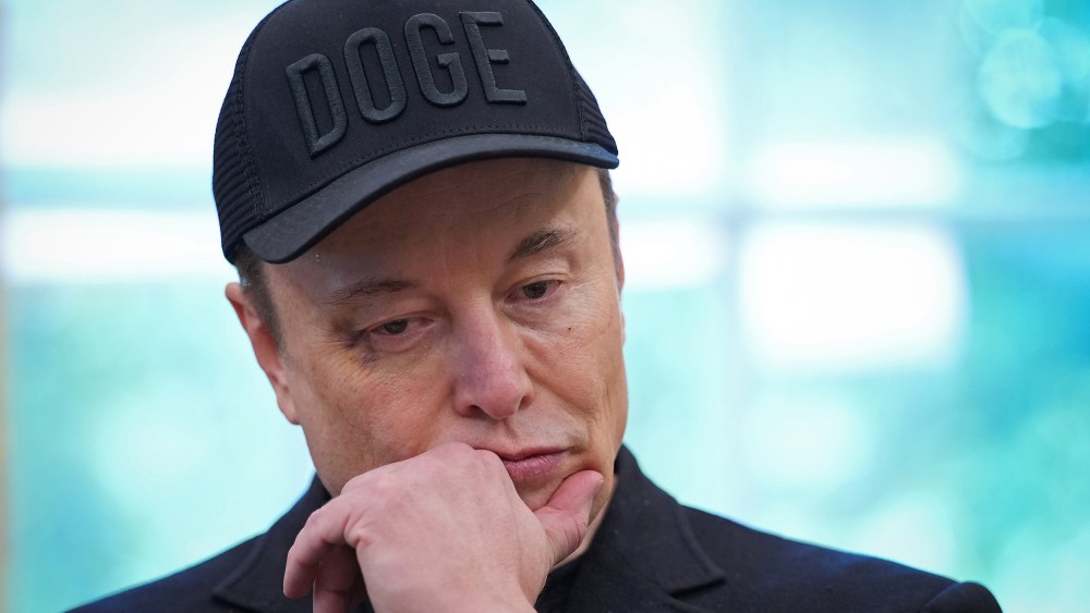 Elon Musk, wearing a black hat that says "DOGE," puts his hand to his chin while looking down.