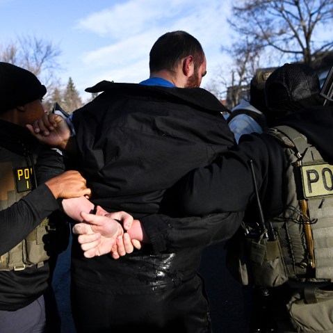 A person with their hands restrained behind their back, surrounded by people in vests labeled "police."