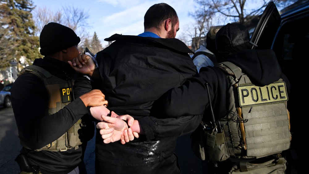 A person with their hands restrained behind their back, surrounded by people in vests labeled "police."