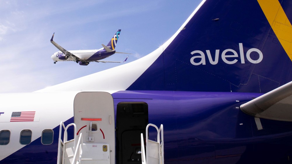 Part of an Avelo airplane is seen in the foreground; an Avelo airplane takes off in the background.