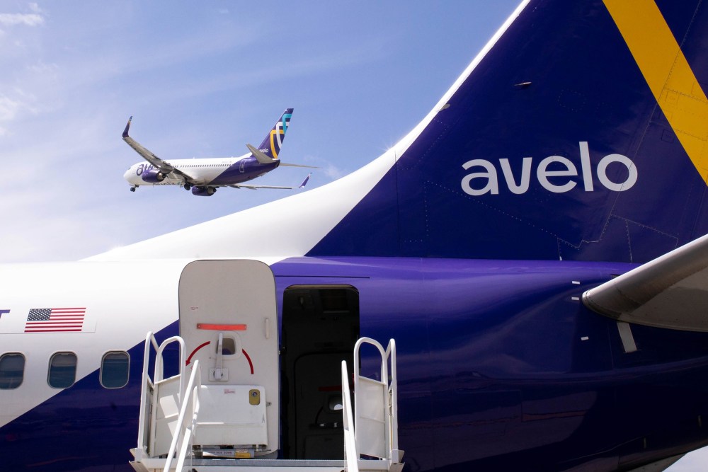 Part of an Avelo airplane is seen in the foreground; an Avelo airplane takes off in the background.