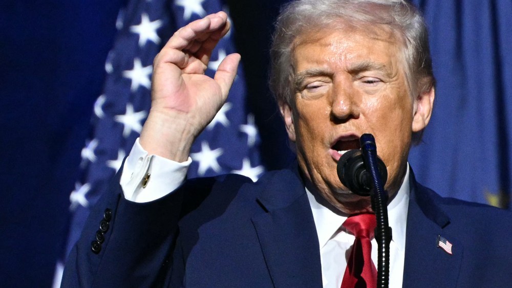 Donald Trump in front of a microphone with his arm raised.