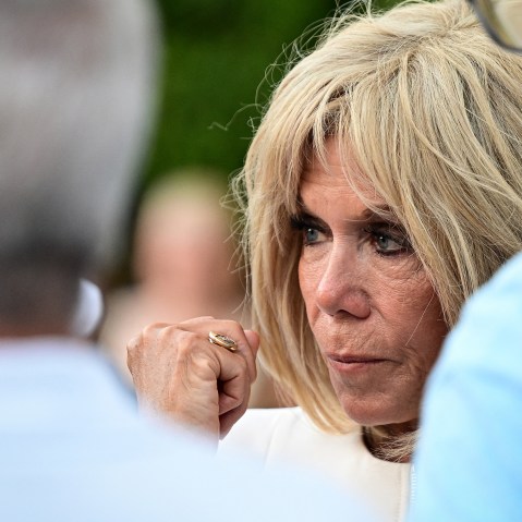 Brigitte Macron, right, looks on while standing with two males. This is a closeup frame.