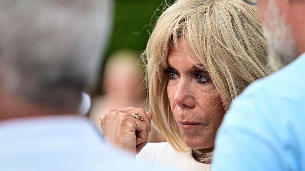 Brigitte Macron, right, looks on while standing with two males. This is a closeup frame.