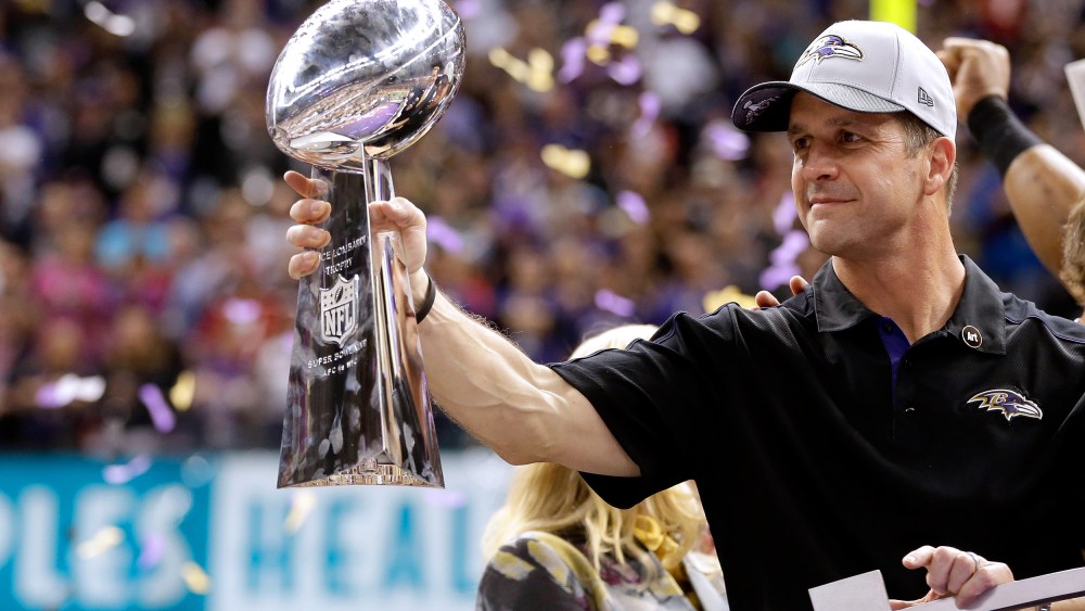 John Harbaugh holds the Vince Lombardi Championship trophy as purple and gold confetti falls in the background.