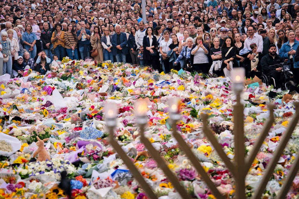 A crowd of people surrounds a large memorial filled with flowers. A menorah stands in the foreground.