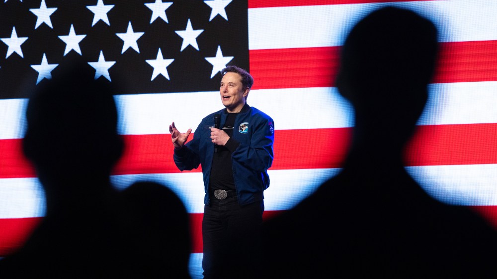 Silhouettes of people watch Elon Musk holding a microphone on a stage backdropped by a digital American flag.