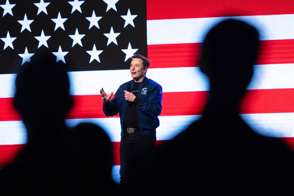 Silhouettes of people watch Elon Musk holding a microphone on a stage backdropped by a digital American flag.