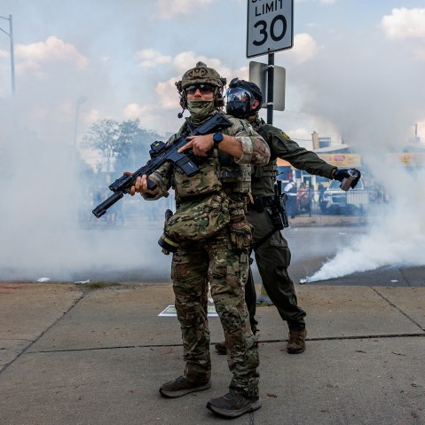 One federal agent stands while another one sprays a foreign agent. There is a protestor wearing a gas mask in the gas cloud.