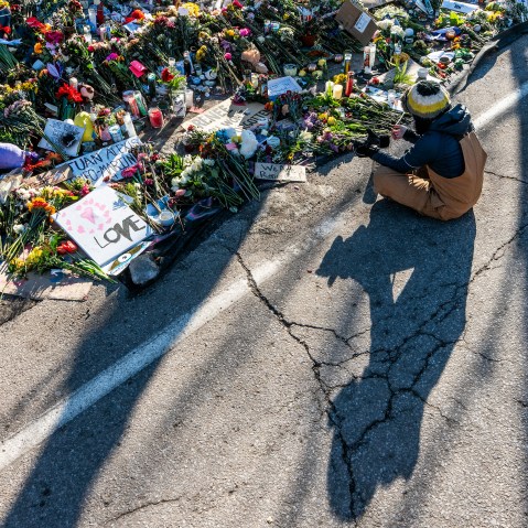 A person sits in the street next to a wealth of flowers, candles, and signs.