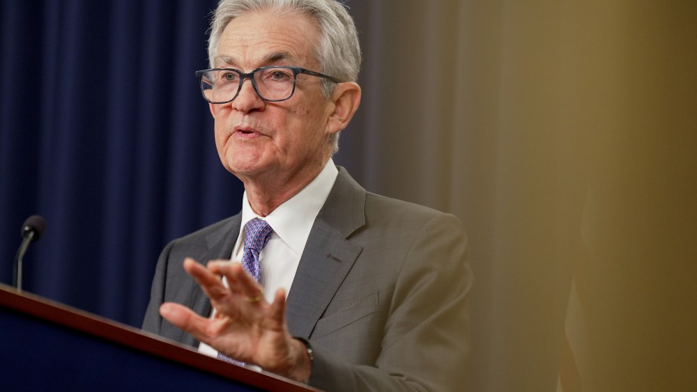 Jerome Powell lifts his left hand while speaking at a podium during a news conference. The photo is shot from below.