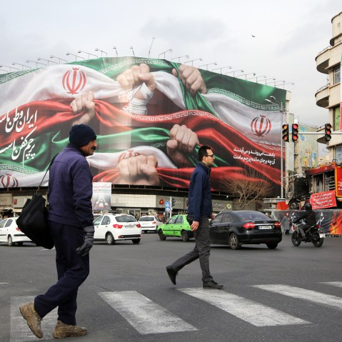 A photo showing daily life in Tehran where two men crossing the street with a big iranian flag seen on a billboard in the background.
