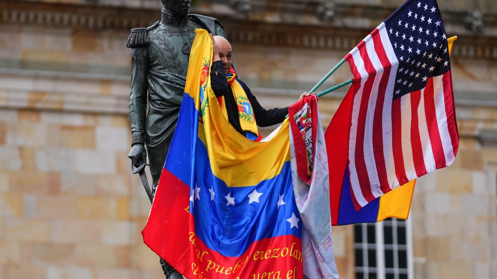 A man waves a venezuelan and american flag while standing a statue in Bogota.