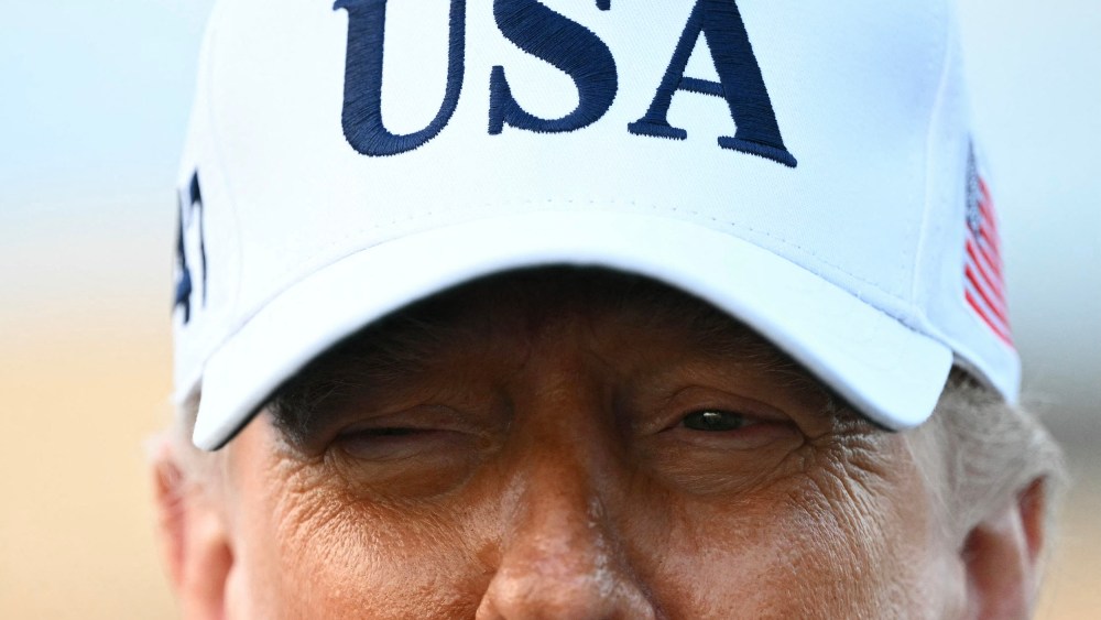 A closeup photo of President Trump shows the upper half of his face wearing a white USA hat.