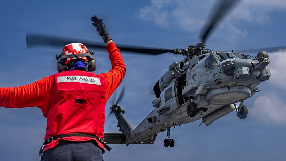 A person wearing red directs a helicopter still in flight.