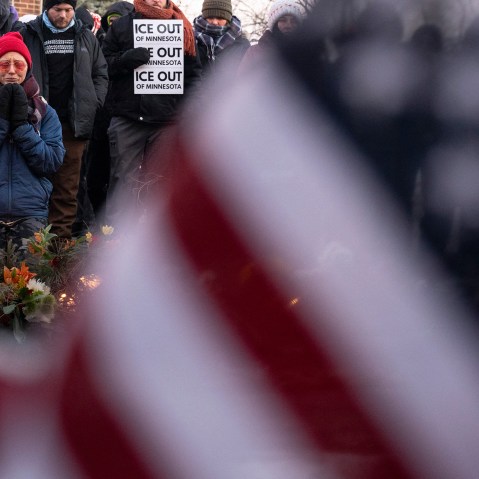 Part of an American flag fills the frame, blurred, in the foreground. On the left of the frame, a person kneels by a memorial of flowers.