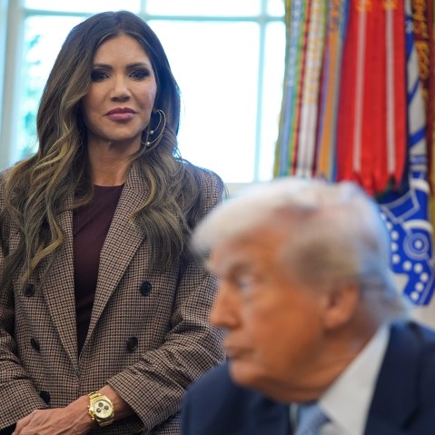 Kristi Noem, left, stands behind Donald Trump, out of focus in the foreground.