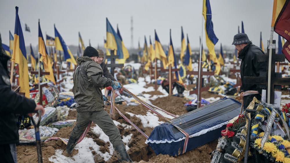Two people pull a casket into a hole in a gravesite covered in crosses and blue and gold Ukrainian flags.