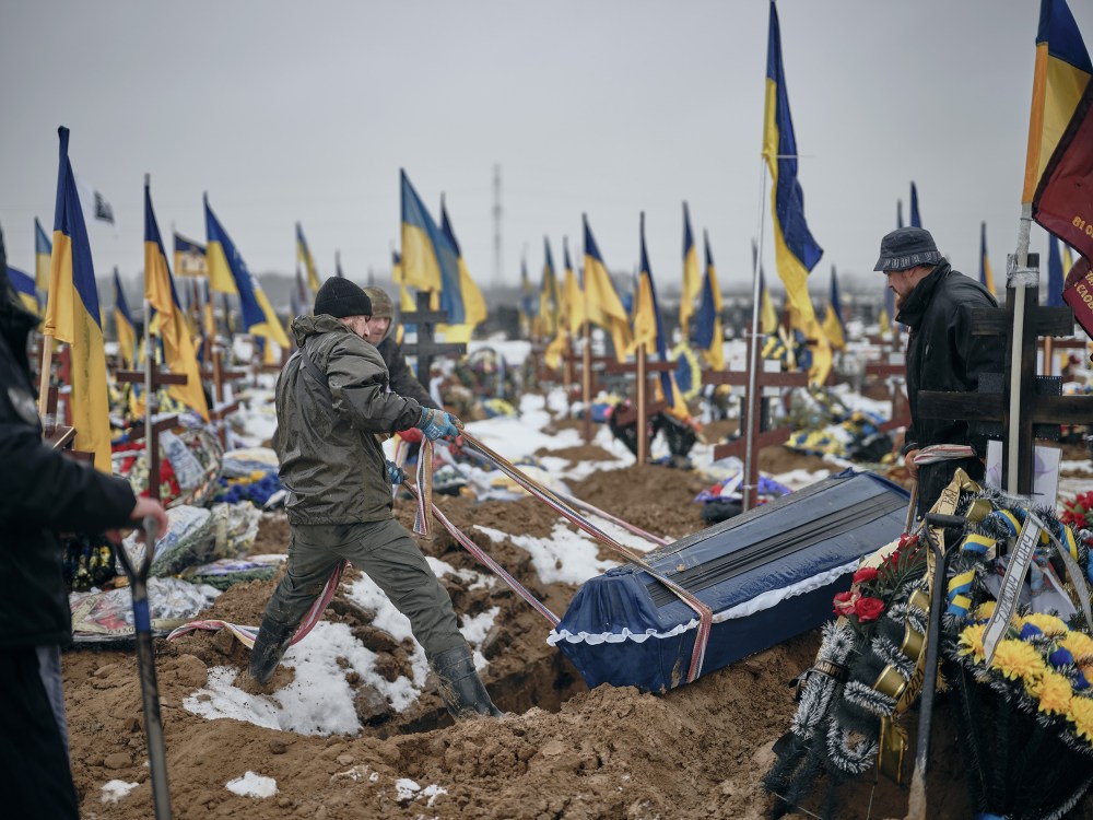 Two people pull a casket into a hole in a gravesite covered in crosses and blue and gold Ukrainian flags.