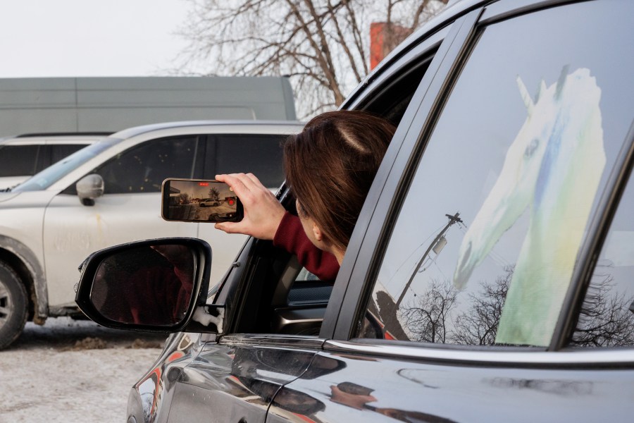 A person in the driver's seat of a car sticks their head and their cell phone out the window to record.