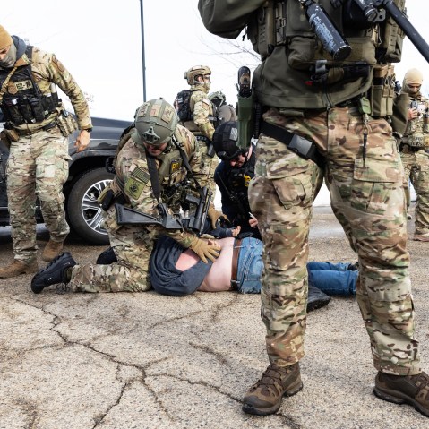 An ICE agent sits on top of a person, who is face down on the ground, and pins the person’s hands behind their back. Other agents stand nearby.