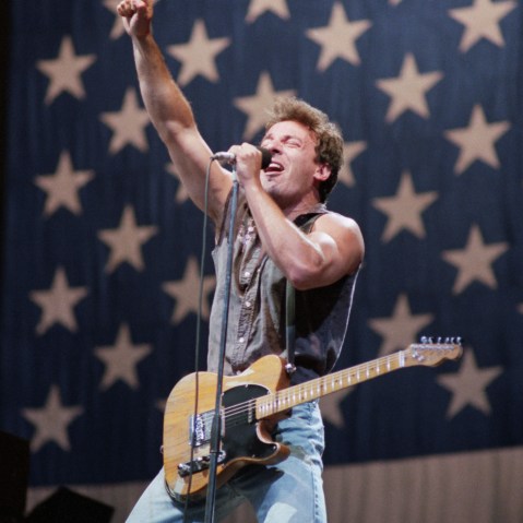 Bruce Springsteen with his guitar on stage, in front of a giant American flag.