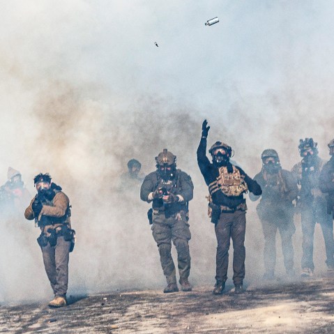 One federal agent, walking through a cloud of tear gas, throws a teargas canister which is now suspended in midair. Other federal agents walk alongside.