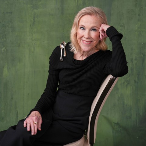 Catherine O'Hara in all black with two spoons on her shoulder. She sits in front of a painted green backdrop.