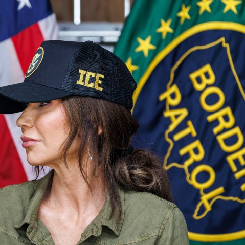 Kristi Noem in an ICE hat in front of an American flag and a U.S. Border Patrol flag.