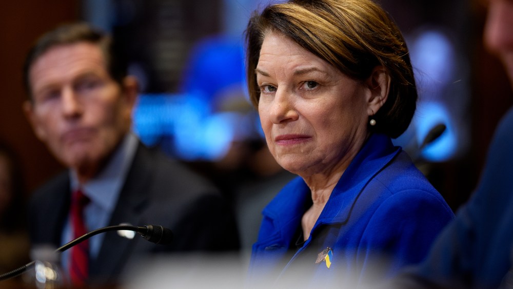 Sen. Amy Klobuchar sitting between other senators while they testify- wearing a bright blue jacket.