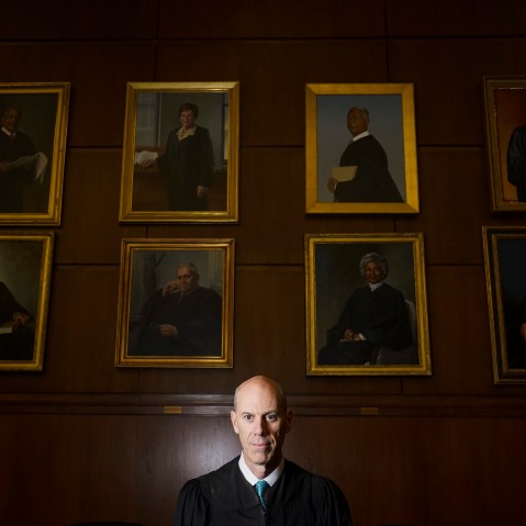 A judge in his robes stands beneath eight gold-framed portraits of other judges.