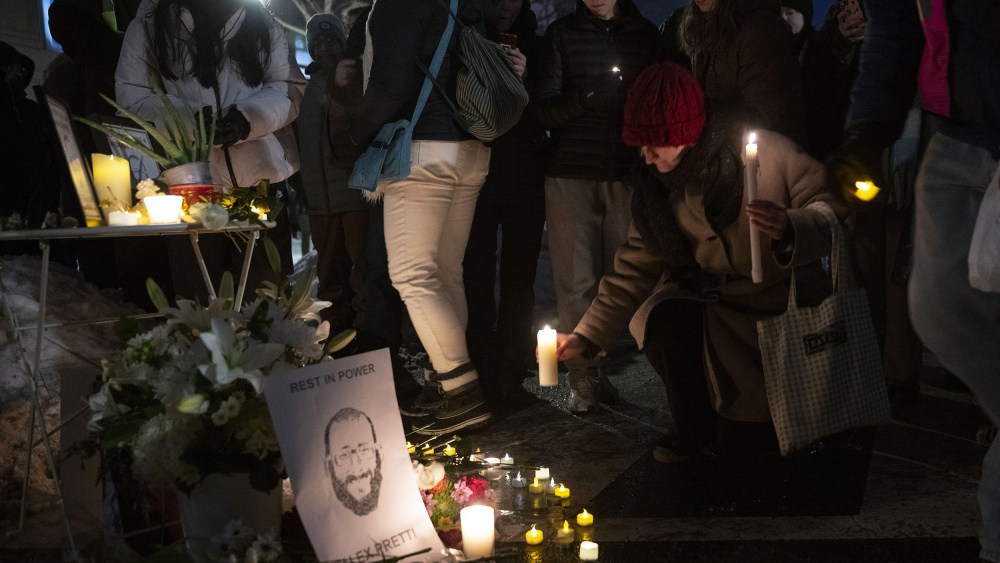 A person kneels down to place a lit candle by a memorial featuring an illustration of Alex Pretti.