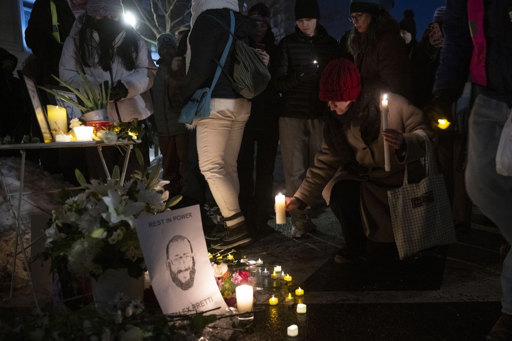A person kneels down to place a lit candle by a memorial featuring an illustration of Alex Pretti.