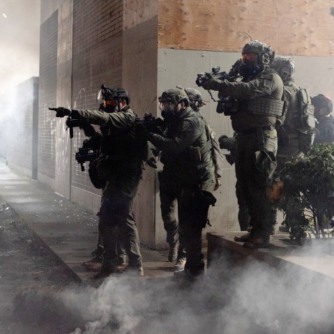 Federal agents are seen in a cloud of smoke in tactical gear, targeting protestors with their weapons.