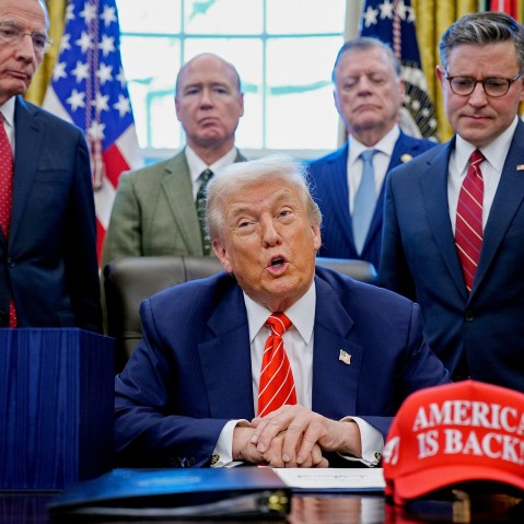 President Donald Trump, center, surrounded by members of the House and Senate in the Oval Office. Out of focus on the desk is a red hat that says "AMERICA IS BACK!"