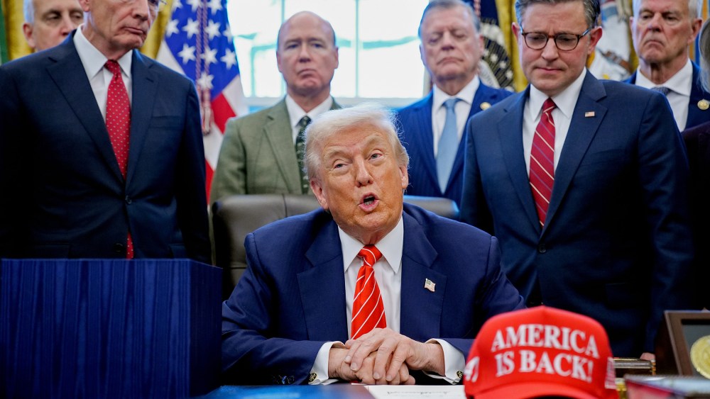 President Donald Trump, center, surrounded by members of the House and Senate in the Oval Office. Out of focus on the desk is a red hat that says "AMERICA IS BACK!"