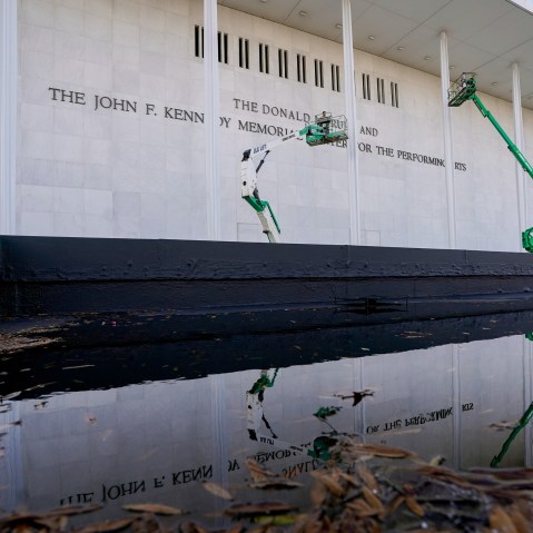 Workers in green construction lifts work on the facade of the Kennedy Center, which is reflected in a puddle.