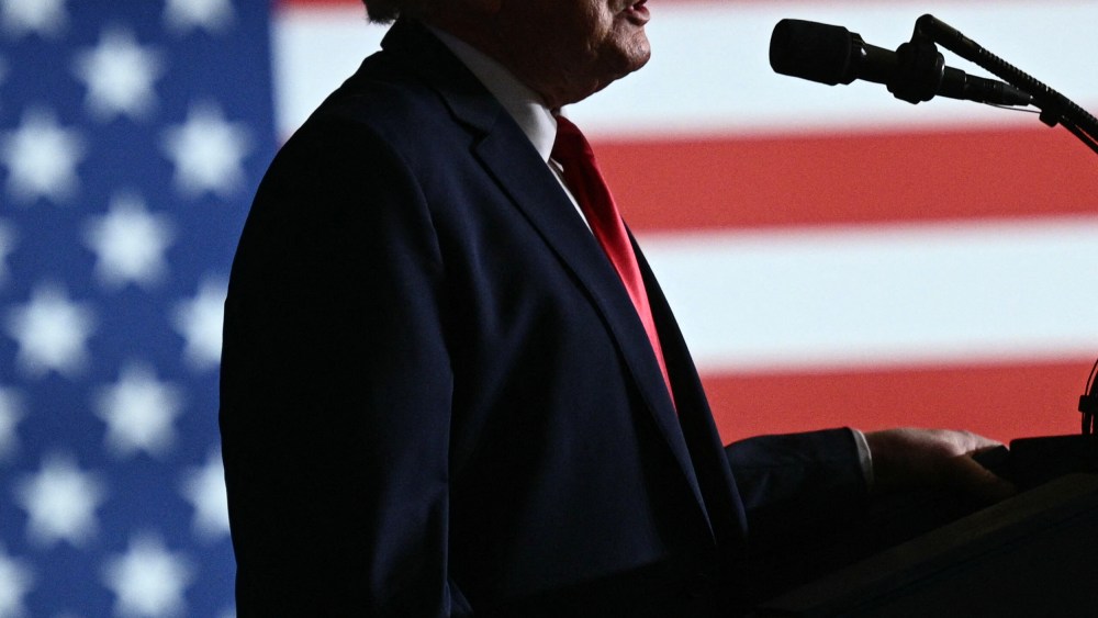 Donald Trump's mouth and torso in profile speaking into a microphone. Behind him, the American flag.