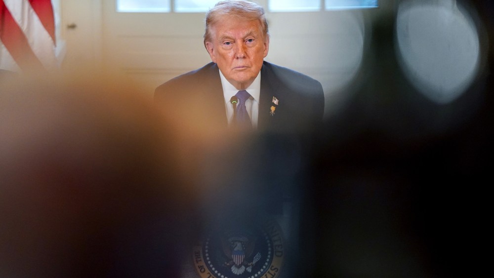 Donald Trump looks on, partially obscured by people in the foreground.