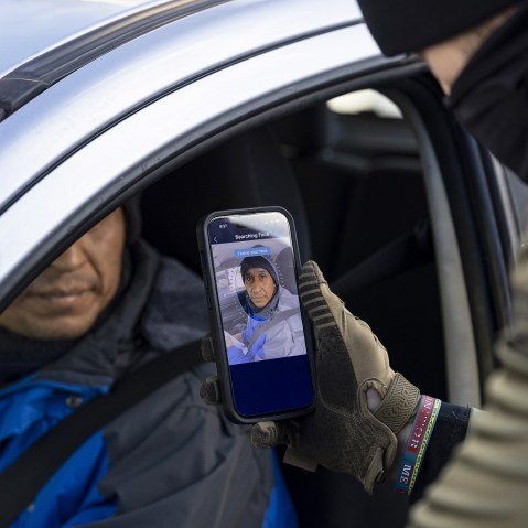 A federal agent holds a phone up to a person in the driver's seat of a car. The phone screen shows a program that is scanning the driver's face.